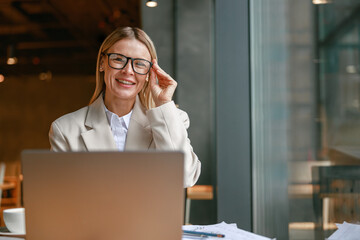 Wall Mural - Businesswoman freelancer working on laptop while sitting in cafe near window and looking at camera