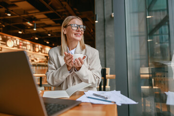 Wall Mural - Attractive businesswoman holding cup of coffee while sitting in cafe during working day