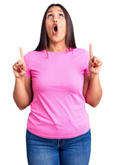 Sticker - Young beautiful brunette woman wearing casual t-shirt amazed and surprised looking up and pointing with fingers and raised arms.