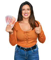 Sticker - Beautiful hispanic woman holding norwegian krone banknotes smiling happy and positive, thumb up doing excellent and approval sign