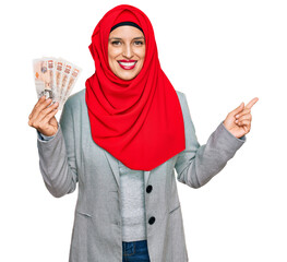 Canvas Print - Beautiful hispanic woman wearing islamic hijab holding 10 pounds banknotes smiling happy pointing with hand and finger to the side