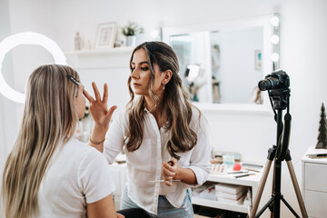 Wall Mural - Happy young woman streaming a beauty makeup vlog from home or workshop. Beautiful online content creator cosmetician applying makeup and explaining some work tools. Vlogging and online channel work.