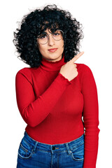 Sticker - Young middle east girl wearing casual clothes and glasses pointing with hand finger to the side showing advertisement, serious and calm face