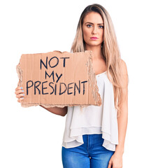 Sticker - Young beautiful blonde woman holding not my president cardboard banner thinking attitude and sober expression looking self confident