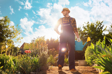 Smiling handsome gardener in uniform posing at kitchen-garden holding a watering can. Bottom view. In the background is a backyard and sunny sky. The concept of gardening and agriculture