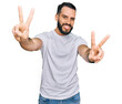 © Krakenimages.com - Young man with beard wearing casual white t shirt smiling with tongue out showing fingers of both hands doing victory sign. number two.