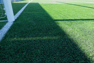 Wall Mural - Artificial turf on football soccer field. Part of soccer goal and green synthetic grass on sport ground with shadow from goal net