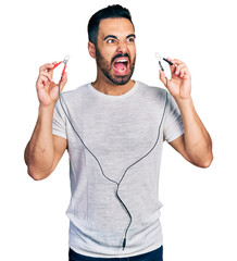 Wall Mural - Young hispanic man with beard holding battery clamps angry and mad screaming frustrated and furious, shouting with anger. rage and aggressive concept.