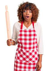 Poster - Young african american woman wearing apron holding rolling pin scared and amazed with open mouth for surprise, disbelief face