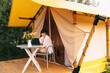 © Ananass - weekend work, Woman starts her morning working on a laptop while sitting near a tent in a glamping site for tourists