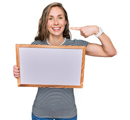 Poster - Young blonde woman holding empty white board pointing finger to one self smiling happy and proud