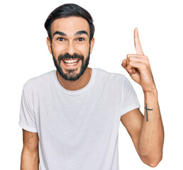 Wall Mural - Young hispanic man wearing casual white t shirt pointing finger up with successful idea. exited and happy. number one.