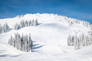 Photo of 3 skiers heading down a widely groomed trail on the mountain from mid-mountain at a ski resort in the Columbia Mountain range in British Columbia, Canada.