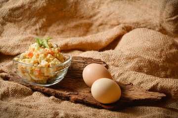 Side view of rustic salad Olivier served in glass bowl on tree bark near two chicken eggs. Country Salad with potato, carrot, green peas, pickled cucumbers, eggs on sackcloth textile background.