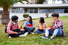Group Portrait Of Happy Teenage Students Holding Notebooks And Smiling Sitting On Courtyard Grass Outdoors. High School Education People. Leisure Time For Young Opponents. Campus University.