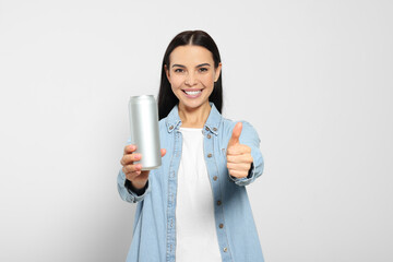 Beautiful happy woman holding beverage can and showing thumbs up on light grey background