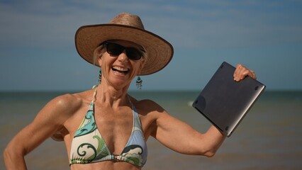 Closeup of pretty happy smiling mature senior woman in hat sunglasses and bikini holding laptop at the beach as digital nomad in tropical place.