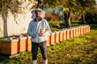 Young experienced beekeeper in protective workwear working at apiary and feeding hives. He is happy and holding a jar of honey while looking at the camera and smiling.