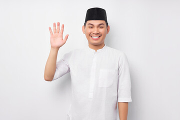 Wall Mural - Smiling friendly young Asian Muslim man wearing Arabic costume tell say hello to camera and waving hand to greet someone isolated over white background. People religious Islamic lifestyle concept