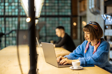 Wall Mural - Young stylish woman works on laptop while sitting by shared table with a coffee drink at modern cafe. Concept of remote work from public place, digital freelance and modern lifestyle