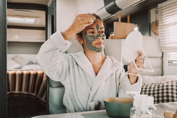 Wall Mural - Portrait of real adult woman applying green mud beauty mask on face using mirror and hands inside a camper van. Alternative lifestyle freedom people travel and nature. Daily routine young female alone