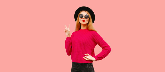 Canvas Print - Portrait of stylish young woman model blowing her red lips sends kiss wearing pink knitted sweater and black round hat isolated on background