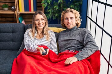 Poster - Young couple sitting on the sofa at home looking positive and happy standing and smiling with a confident smile showing teeth