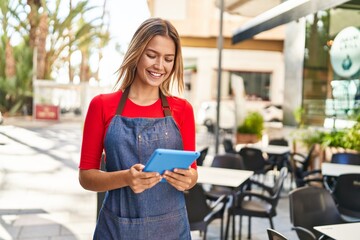 Wall Mural - Young hispanic woman waitress smiling confident using touchpad at coffee shop terrace