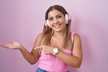 Canvas Print - Young blonde woman listening to music using headphones amazed and smiling to the camera while presenting with hand and pointing with finger.