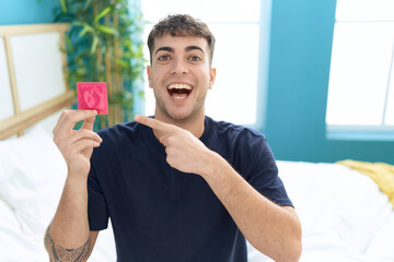 Young hispanic man sitting on the bed holding condom smiling happy pointing with hand and finger