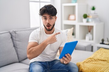 Sticker - Hispanic man with beard using touchpad sitting on the sofa surprised pointing with finger to the side, open mouth amazed expression.