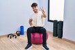 Hispanic man with beard sitting on pilate balls at yoga room smiling looking to the camera showing fingers doing victory sign. number two.