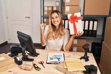 Poster - Young blonde woman working at small business ecommerce holding gift screaming proud, celebrating victory and success very excited with raised arm