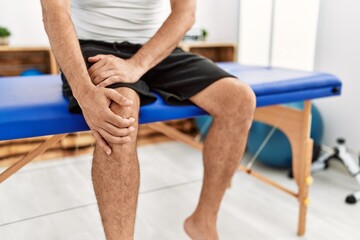 Poster - Middle age grey-haired man patient suffering for knee injury sitting on massage table at rehab clinic