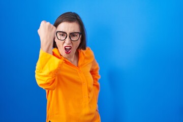 Sticker - Middle age hispanic woman wearing glasses standing over blue background angry and mad raising fist frustrated and furious while shouting with anger. rage and aggressive concept.