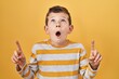 Young caucasian kid standing over yellow background amazed and surprised looking up and pointing with fingers and raised arms.