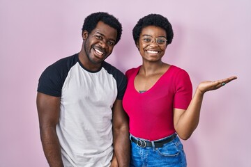 Sticker - Young african american couple standing over pink background smiling cheerful presenting and pointing with palm of hand looking at the camera.