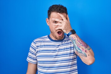 Canvas Print - Young hispanic man standing over blue background peeking in shock covering face and eyes with hand, looking through fingers with embarrassed expression.