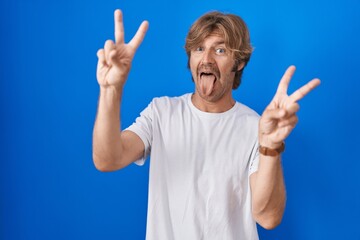 Wall Mural - Middle age man standing over blue background smiling with tongue out showing fingers of both hands doing victory sign. number two.