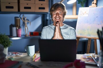 Canvas Print - Middle age man sitting at art studio with laptop at night praying with hands together asking for forgiveness smiling confident.