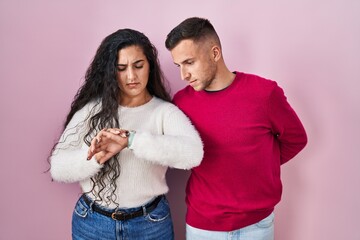 Wall Mural - Young hispanic couple standing over pink background checking the time on wrist watch, relaxed and confident