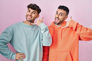 Wall Mural - Young hispanic gay couple standing over pink background smiling doing phone gesture with hand and fingers like talking on the telephone. communicating concepts.