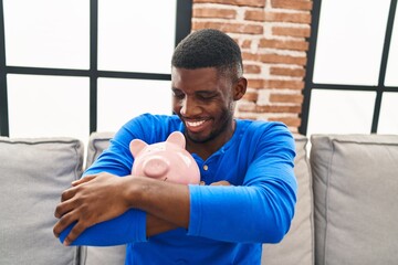 Wall Mural - Young african american man hugging piggy bank sitting on sofa at home