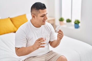 Sticker - Young latin man smiling confident holding pills and glass of water at bedroom