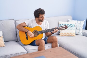 Canvas Print - Young hispanic man playing classical guitar sitting on sofa at home