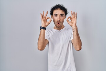 Sticker - Hispanic man standing over isolated background looking surprised and shocked doing ok approval symbol with fingers. crazy expression