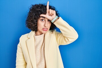 Poster - Young brunette woman with curly hair standing over blue background making fun of people with fingers on forehead doing loser gesture mocking and insulting.