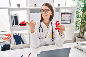 Poster - Young caucasian doctor woman holding support red ribbon pointing thumb up to the side smiling happy with open mouth