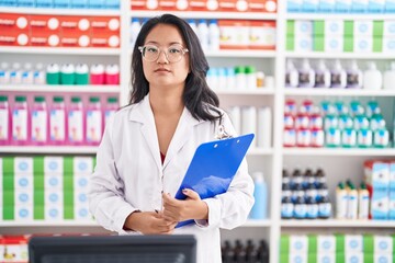 Canvas Print - Asian young woman working at pharmacy drugstore holding clipboard thinking attitude and sober expression looking self confident