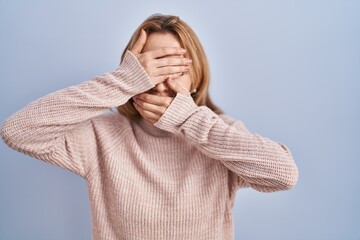 Wall Mural - Hispanic woman standing over blue background covering eyes and mouth with hands, surprised and shocked. hiding emotion